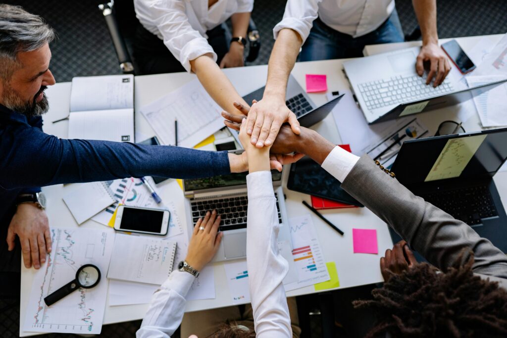 High-angle shot of diverse hands together over a desk with laptops and paperwork, symbolizing teamwork.